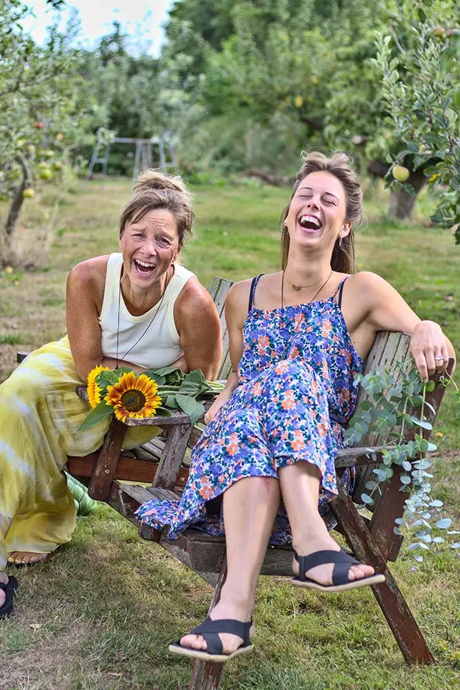 Ingrid en Anouk zitten lachend naast elkaar op een houten bank in een boomgaard, met een zonnebloem en eucalyptustakken naast zich
