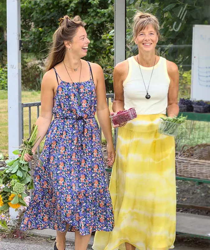 Ingrid en Anouk lopen lachend naast elkaar langs een kraam met verse groenten, met een bos bloemen, een bakje frambozen en sperziebonen in hun handen