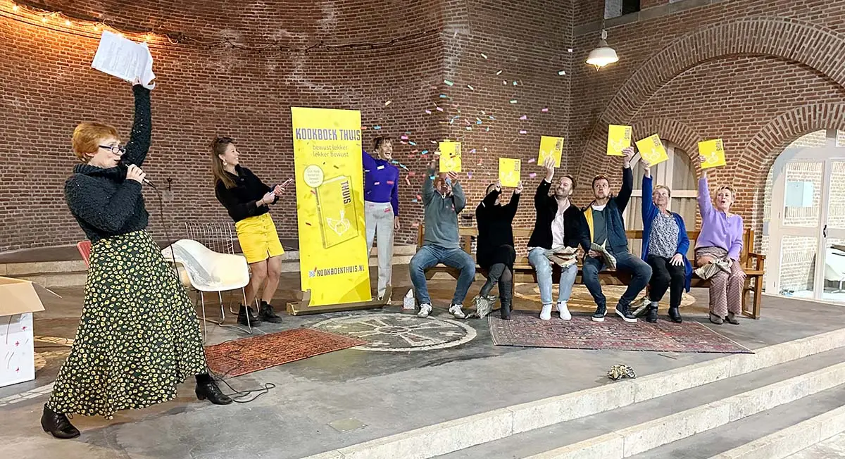 De boekpresentatie van kookboek THUIS in een zaal met bakstenen gewelven: bezoekers houden lachend het gele boek omhoog terwijl er confetti door de lucht vliegt