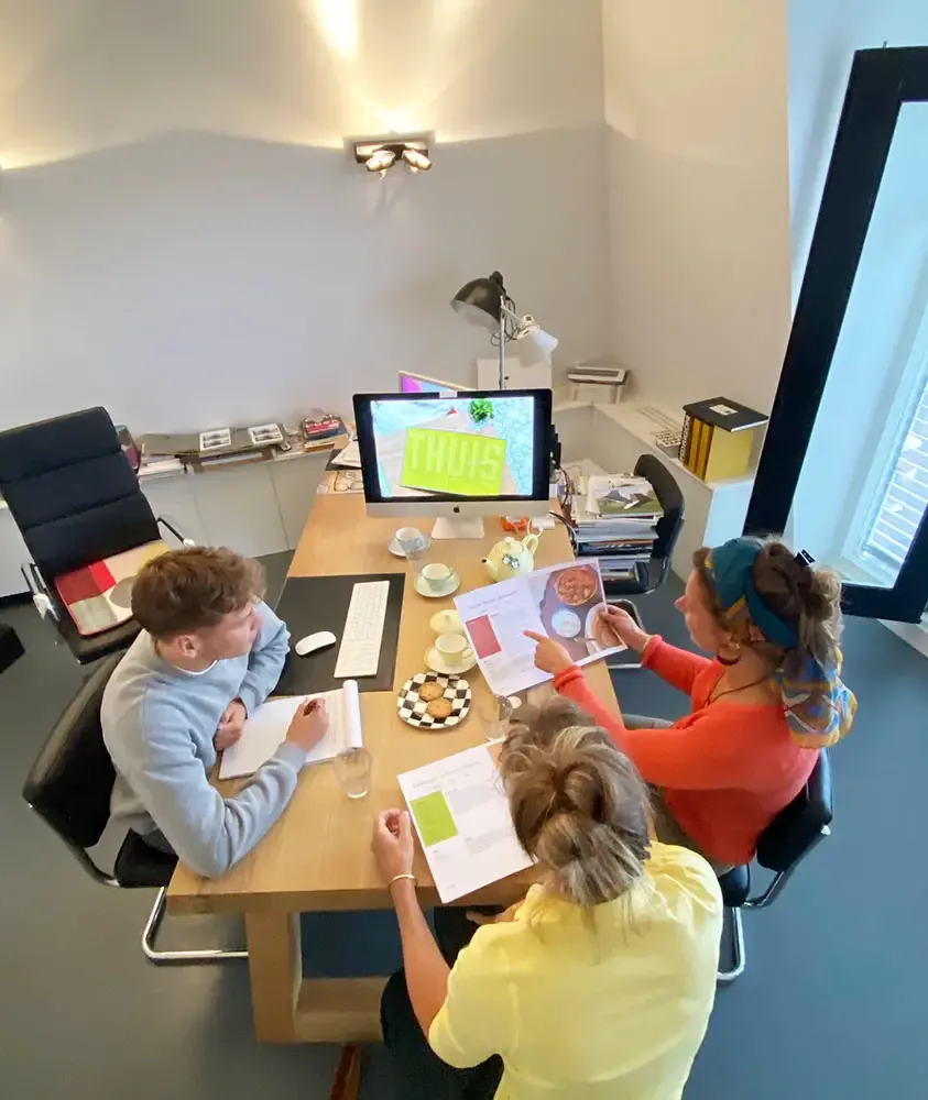 Van boven gezien: drie mensen aan een vergadertafel bekijken uitgeprinte proefpagina’s van een kookboek en maken aantekeningen, met op het beeldscherm de titel THUIS